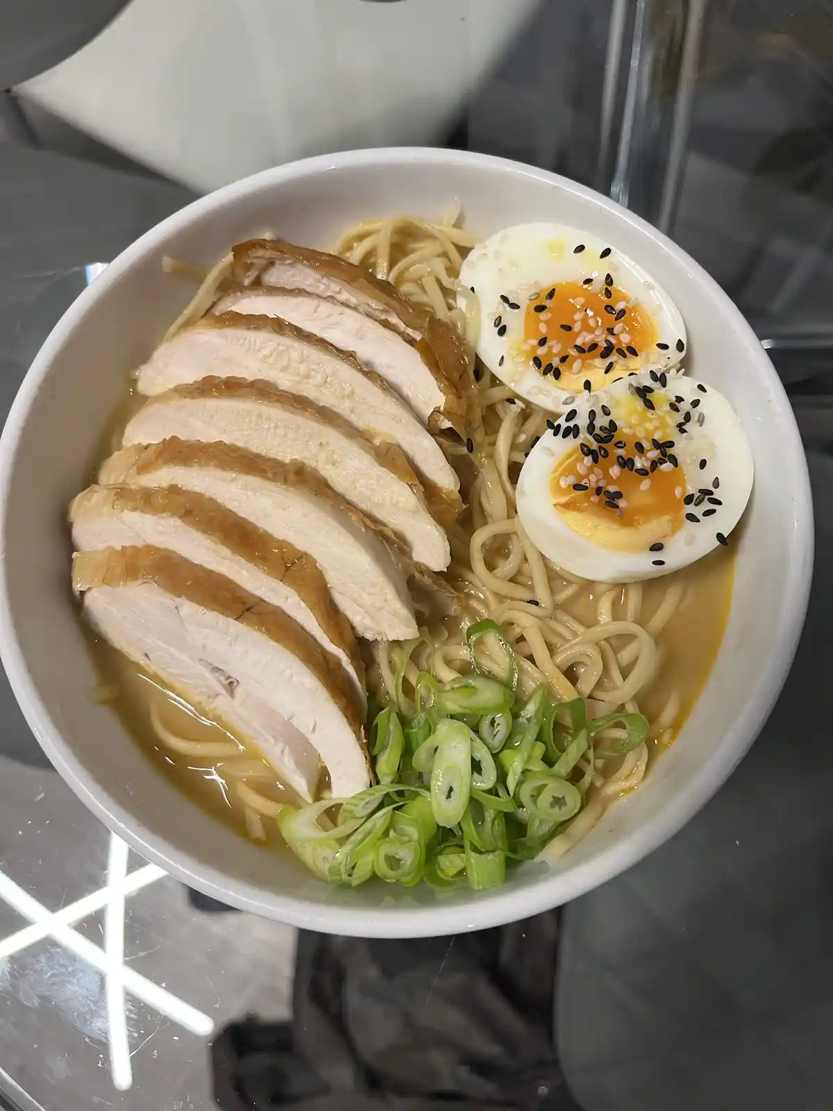 A top-down photo of a white bowl filled with ramen noodles, topped with sliced chicken, two halves of a soft-boiled egg…
