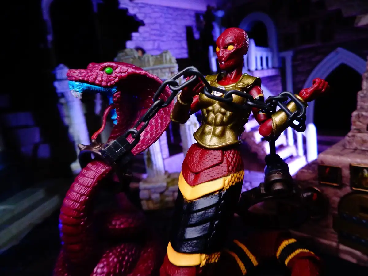 A red-skinned action figure with glowing yellow eyes and gold armor stands holding a large black chain attached to a red…