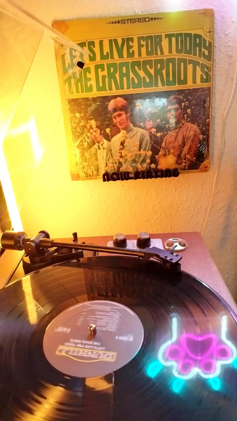 A vinyl record by The Grassroots titled 'Let's Live For Today' spins on a turntable, with the album cover mounted on the…
