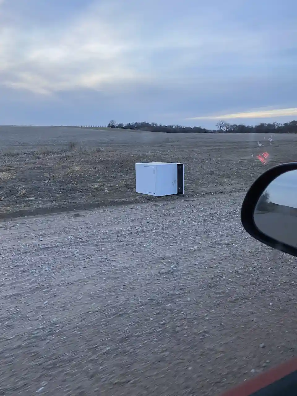 A white microwave oven lies on its side in the middle of a barren, gravelly field, viewed from inside a moving car with the…