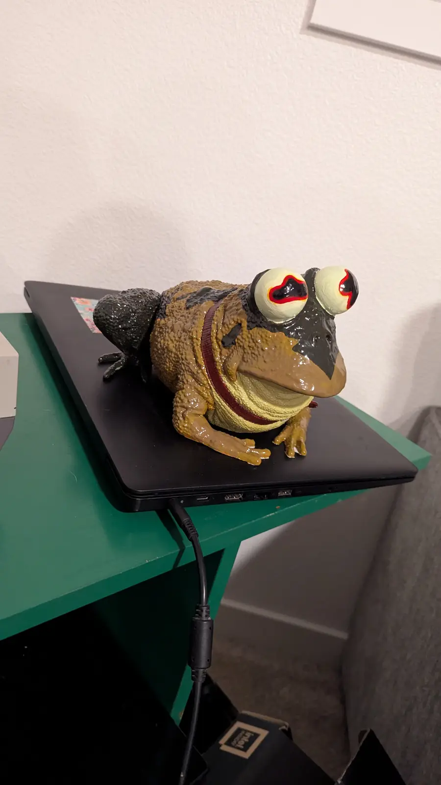 A large, textured frog figurine with oversized, cartoonish eyes sits atop a closed black laptop on a green table. The frog…