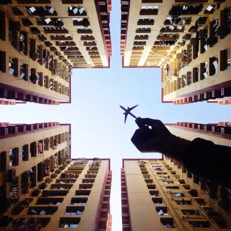 A low-angle photo shows a hand holding a small airplane model against a bright blue sky, framed by four tall apartment…