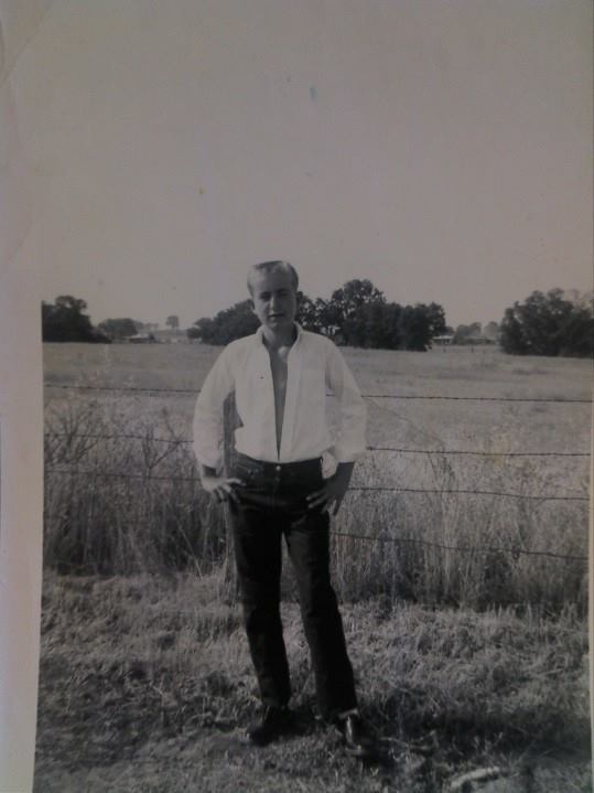 A black-and-white photo shows a young man standing outdoors in a field, hands on hips, in front of a barbed wire fence. He…
