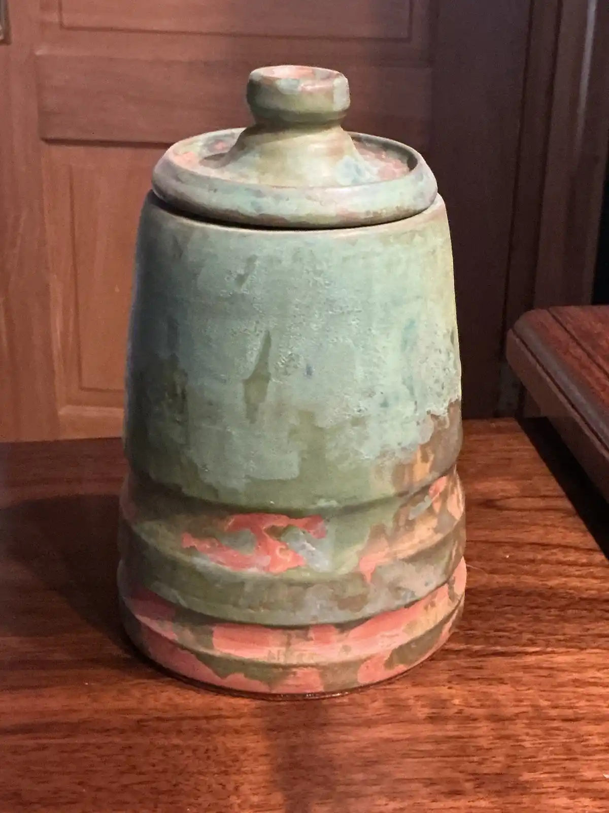 A ceramic jar with a lid sits on a wooden surface, its greenish glaze heavily chipped and worn, revealing patches of…
