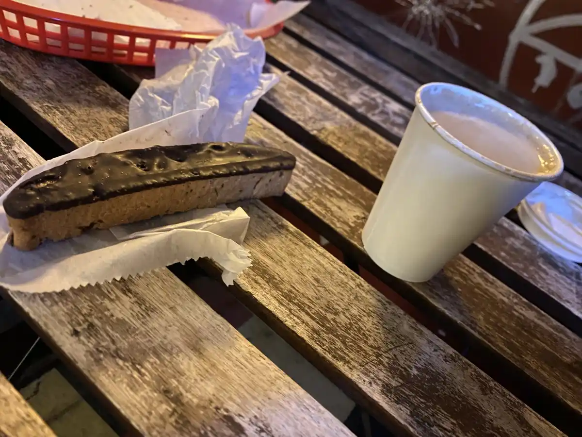 A chocolate-covered biscotti sits on white paper next to a paper cup of coffee on a weathered wooden slatted table. In the…