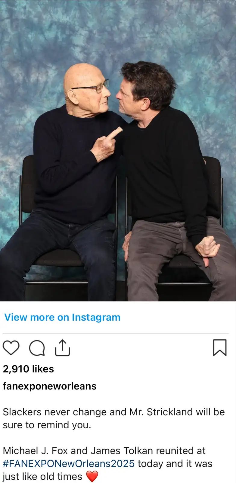 Two men, Michael J. Fox and James Tolkan, sit facing each other in chairs against a blue textured backdrop. The older man…