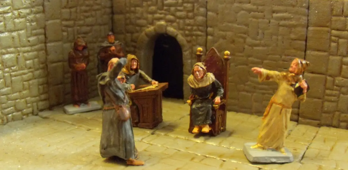 A diorama of medieval figures in a stone-walled room, featuring a seated king on a throne, a man in a grey robe gesturing…
