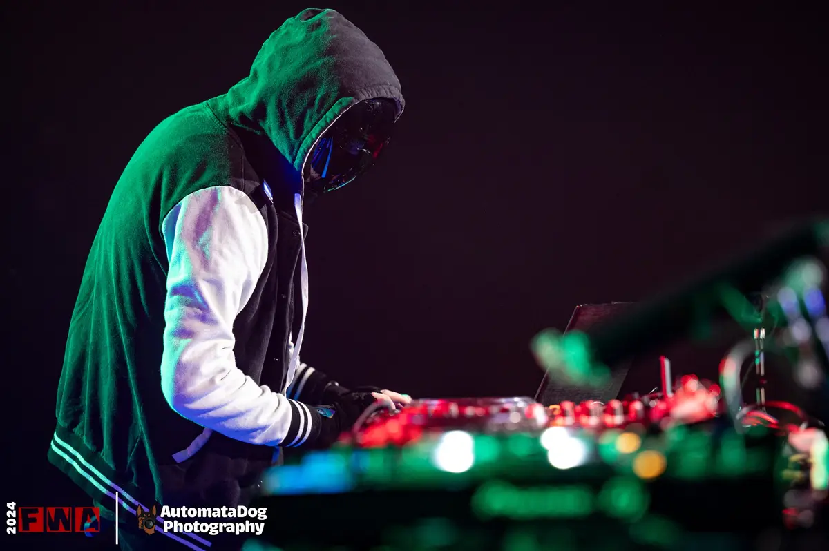 A person wearing a green hoodie and a black mask is leaning over a glowing red and green DJ controller. The image has a dark…