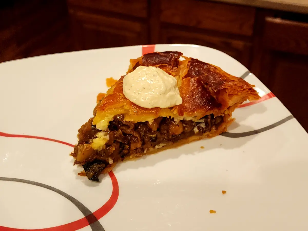 A close-up photo shows a slice of savory pie with a golden-brown, flaky crust and a filling of meat and vegetables, topped…