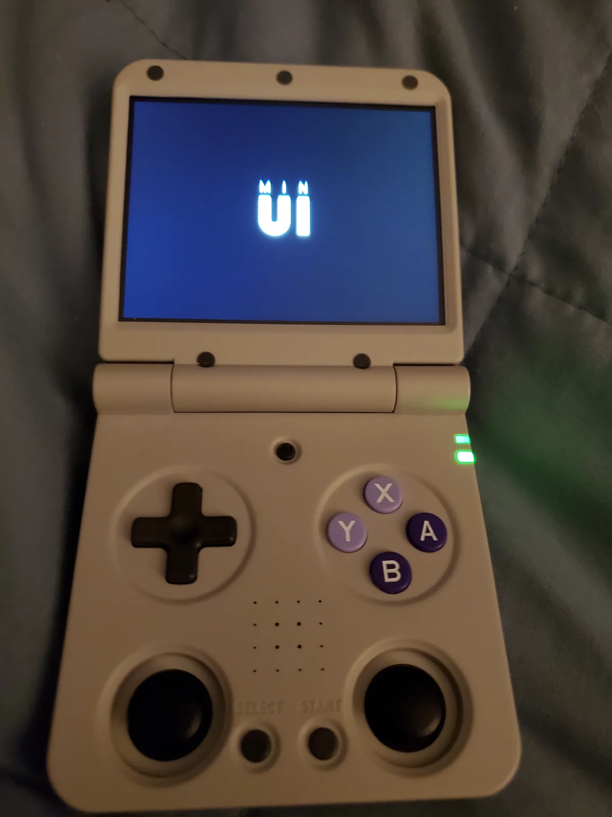A beige handheld gaming device, resembling a Nintendo Game Boy Advance, is open and powered on, displaying the text 'MIN UI'…