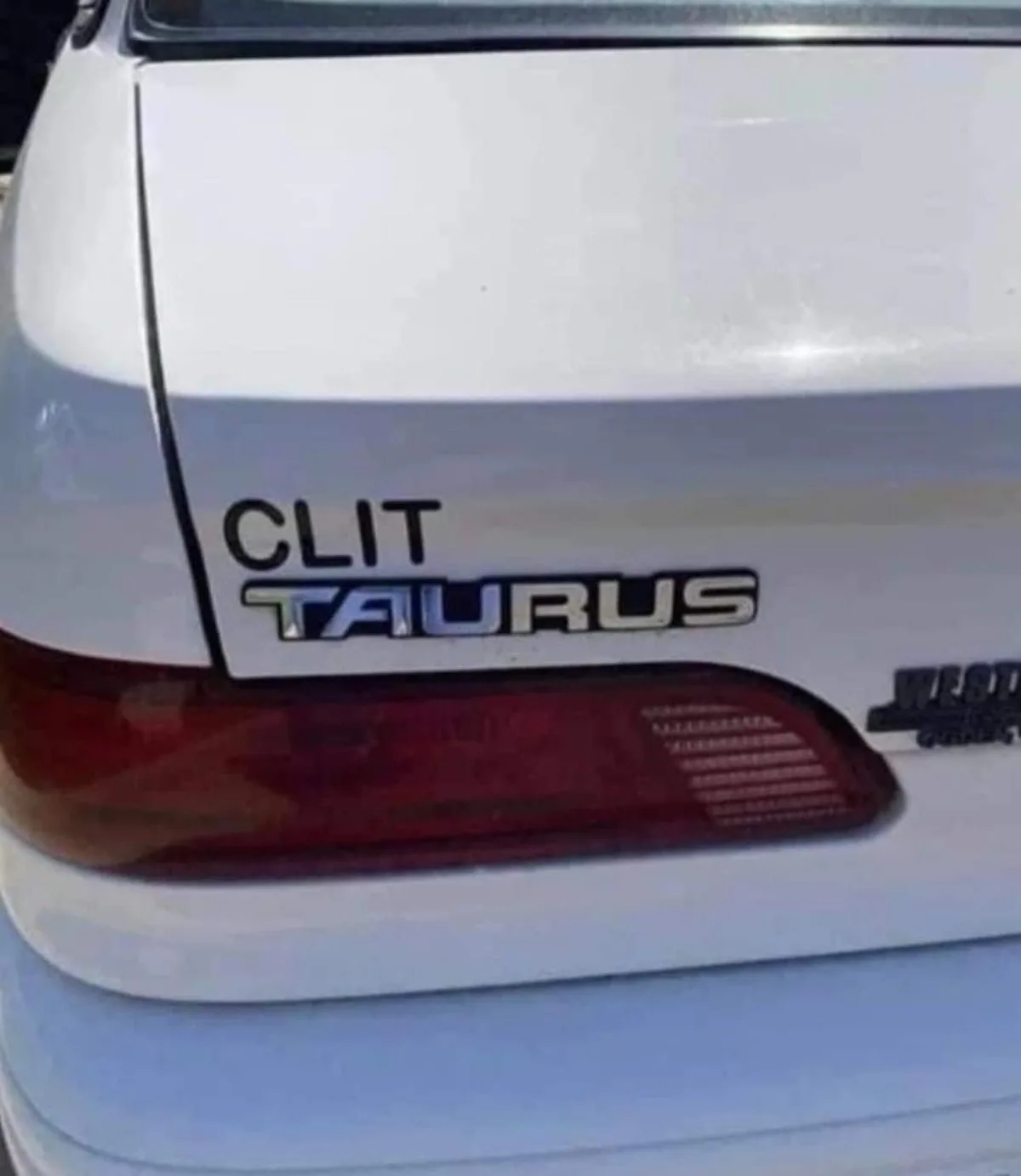 A close-up of the rear of a white car, showing a modified emblem where the word 'CLIT' is added above the official 'TAURUS'…