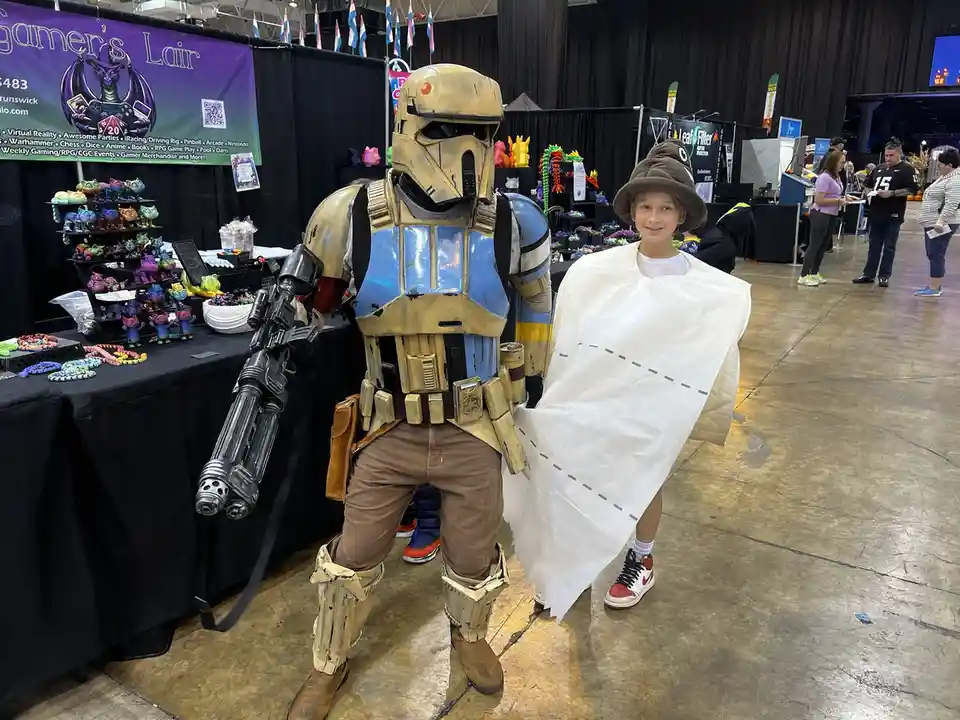 A person in a detailed Star Wars clone trooper costume stands beside a young girl wearing a brown poop emoji hat and a white…