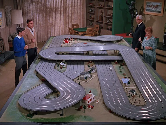 A still from a 1960s TV show shows four people standing around a large, elaborate model slot car track with multiple loops…