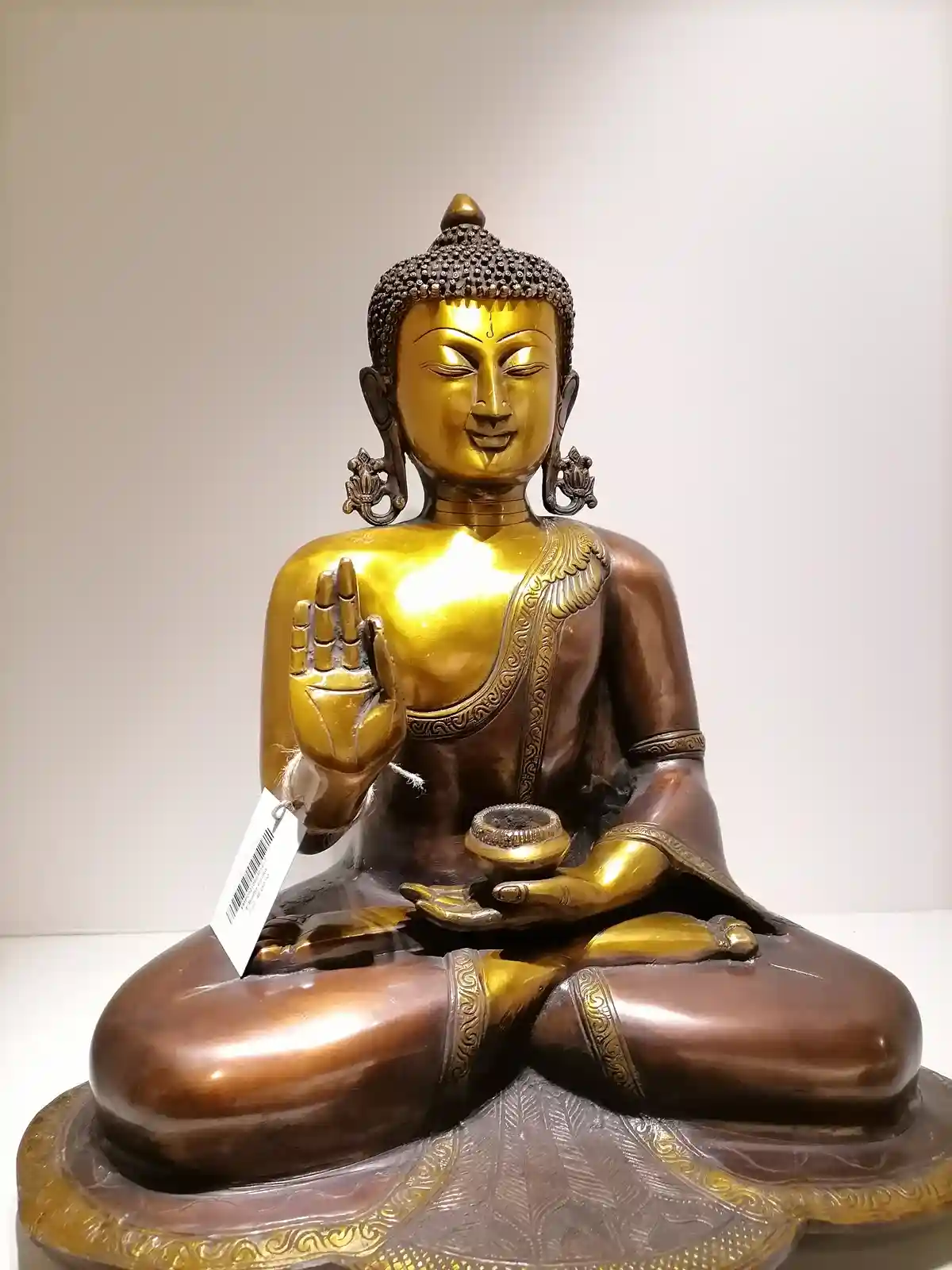 A golden Buddha statue sits in a meditative pose, holding a small bowl in one hand and a price tag attached to the other…