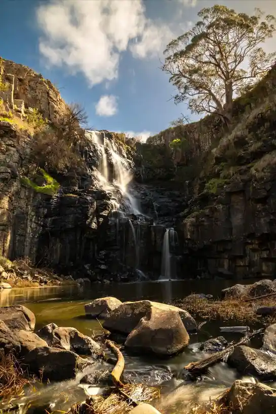 A serene, long-exposure photograph of a waterfall cascading down dark, layered rock cliffs into a calm pool surrounded by…