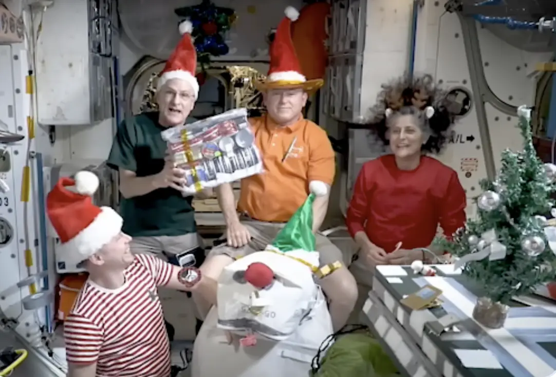 four astronauts in a cramped space station module, all wearing red santa hats and festive attire, celebrate christmas with a…