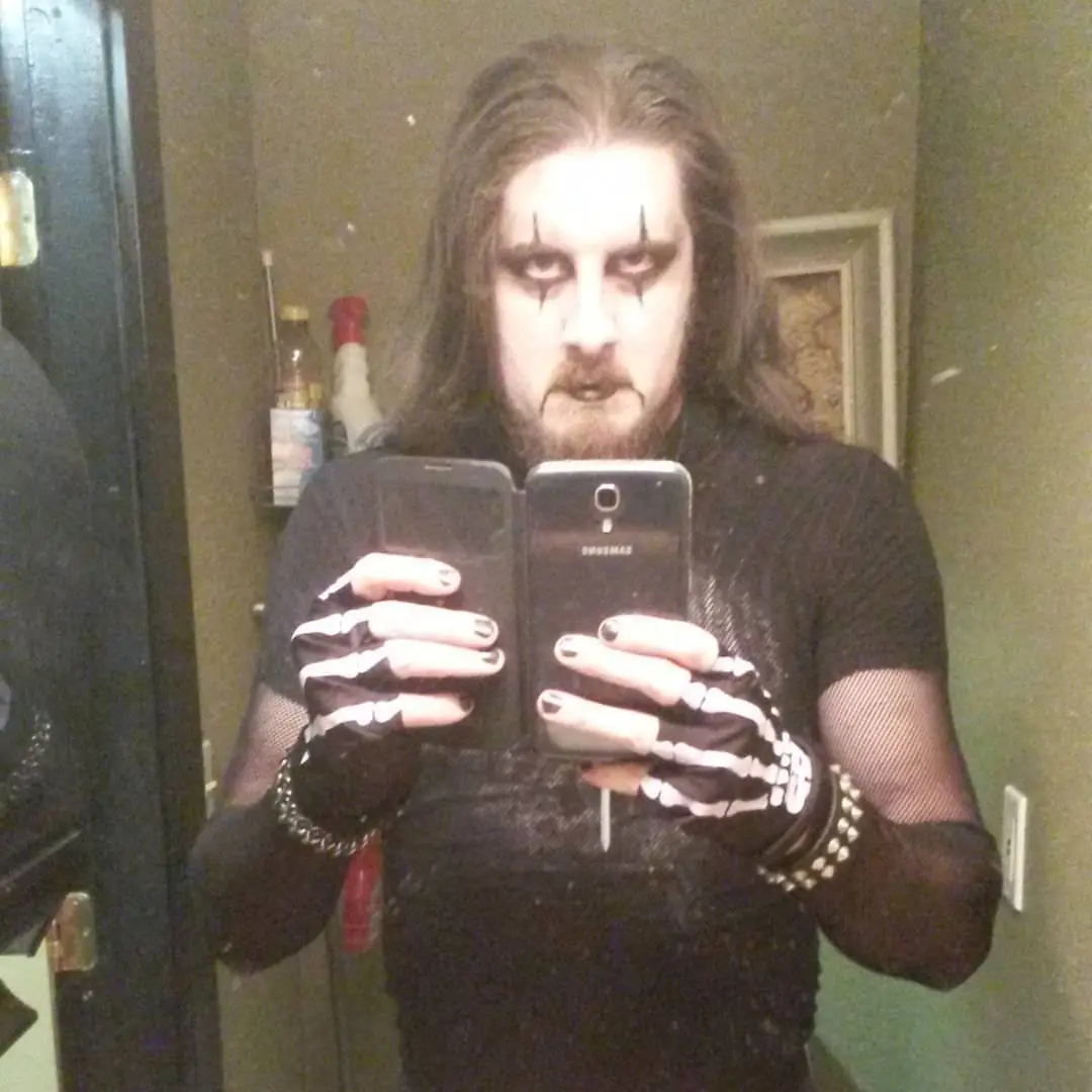 A person with long dark hair, black face paint with sharp white markings, and a beard takes a mirror selfie holding two…