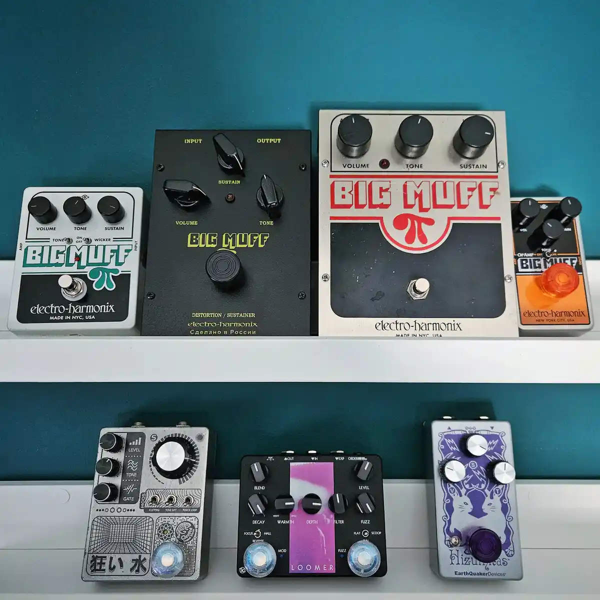 Seven guitar effect pedals are arranged on two white shelves against a teal wall. Four pedals in the top row are variations…