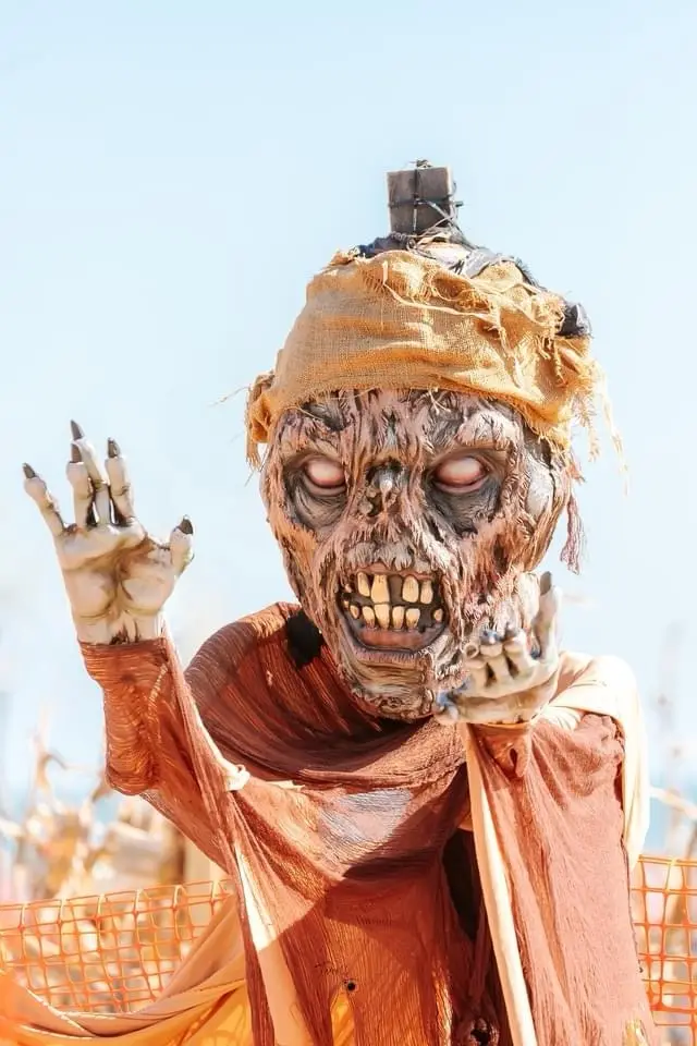 A person in a detailed, gruesome zombie costume with tattered orange fabric, a burlap head wrap, and clawed hands raised in…