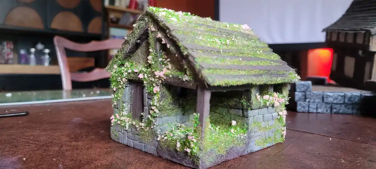 A miniature model of a rustic cottage with mossy roof and walls covered in pink and white flowers sits on a wooden table…