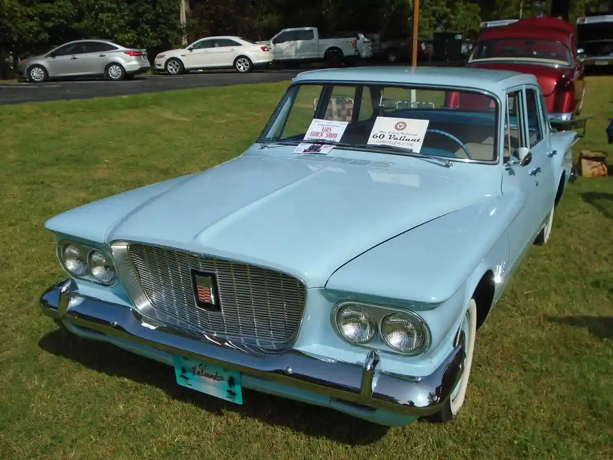 A light blue 1960 Chrysler Valiant sedan is parked on grass at a car show, with a sign in the windshield reading '60 Valiant…