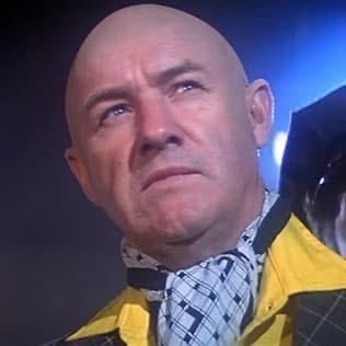 A bald man with a serious, slightly disgusted expression looks upward and to his left. He wears a yellow jacket over a plaid…