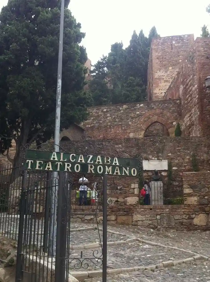 A photo of an ornate metal gate with a green sign that reads 'ALCAZABA TEATRO ROMANO' in white letters. Behind the gate are…