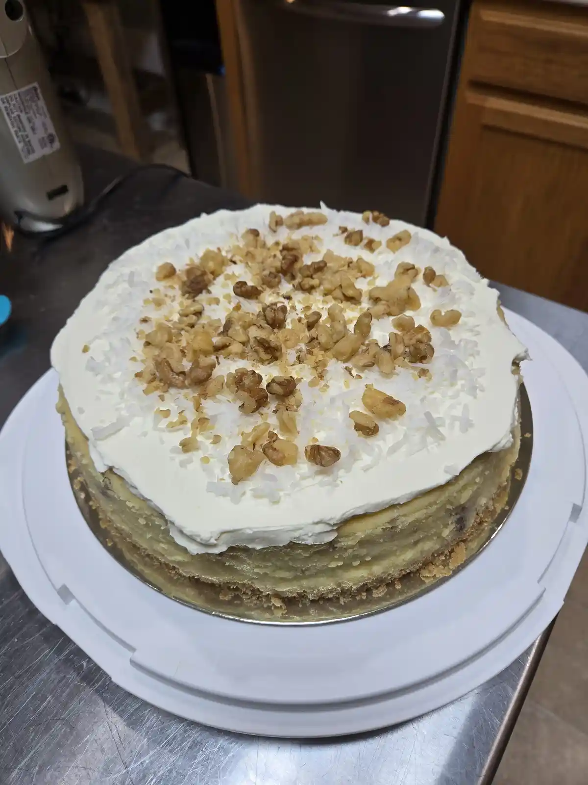 A round cake with white frosting, shredded coconut, and chopped walnuts sits on a white cake board on a stainless steel…
