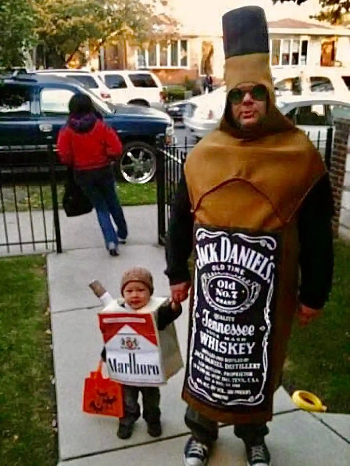 An adult dressed in a full-body Jack Daniel's whiskey bottle costume stands on a sidewalk holding a small child's hand. The…