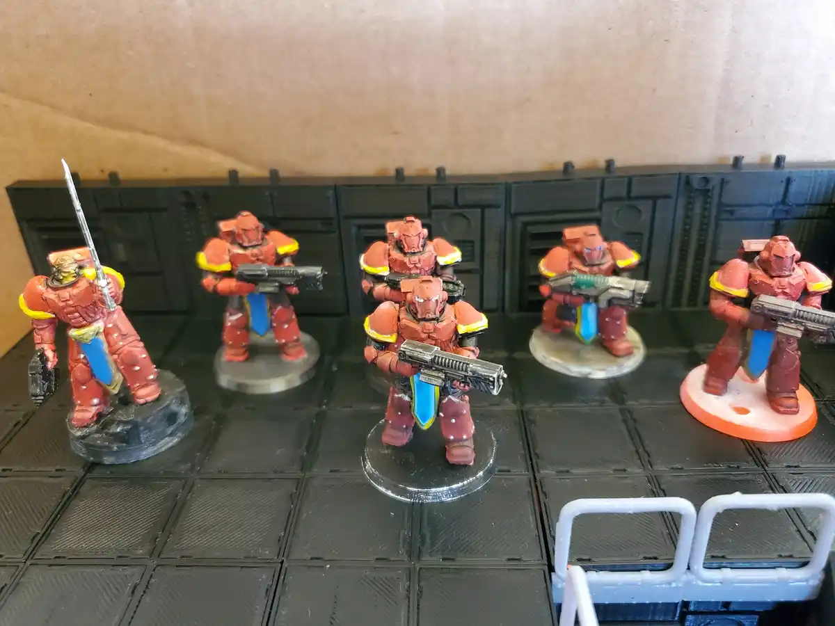Six painted miniature figures in red armor with yellow and blue accents stand on a black grid battlefield. Each figure holds…