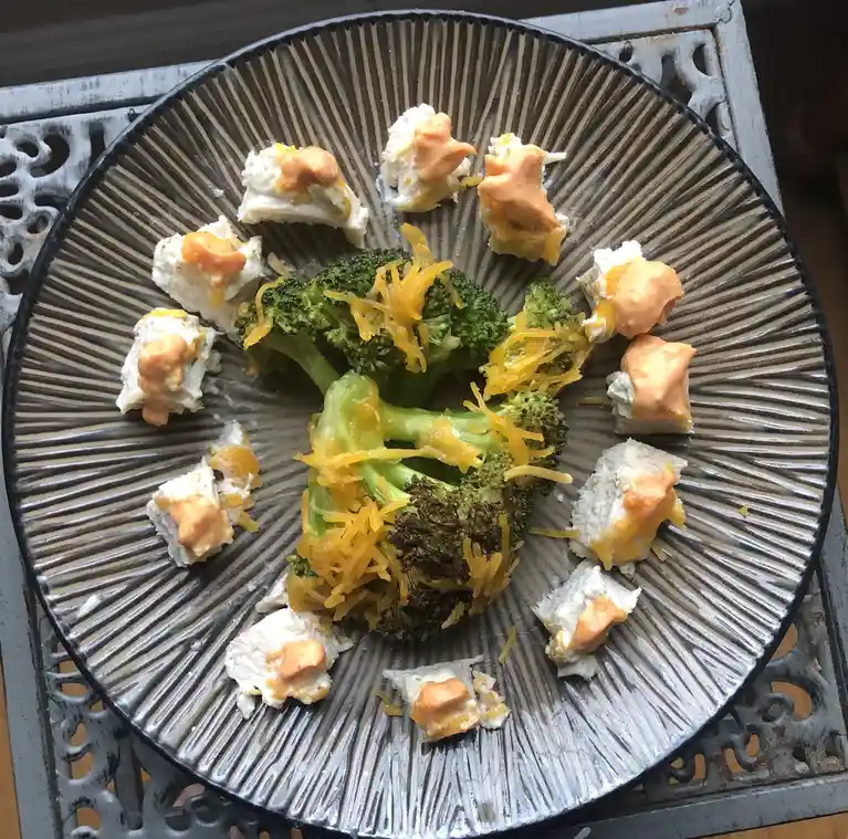 A plate of food features a central floret of broccoli topped with shredded cheese, surrounded by small pieces of what…