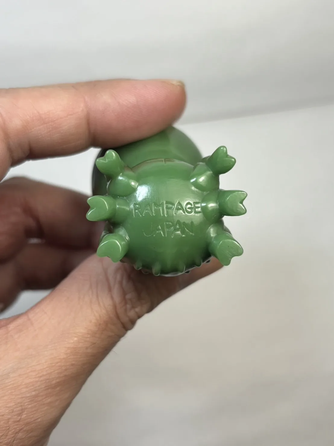 A hand holds a small, green plastic toy figure, showing its underside which has eight claw-like feet and the embossed text…