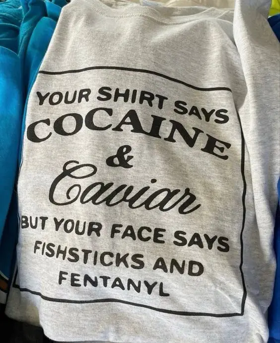 A light gray t-shirt with black text in a label-style layout, reading 'YOUR SHIRT SAYS COCAINE & Caviar BUT YOUR FACE SAYS…