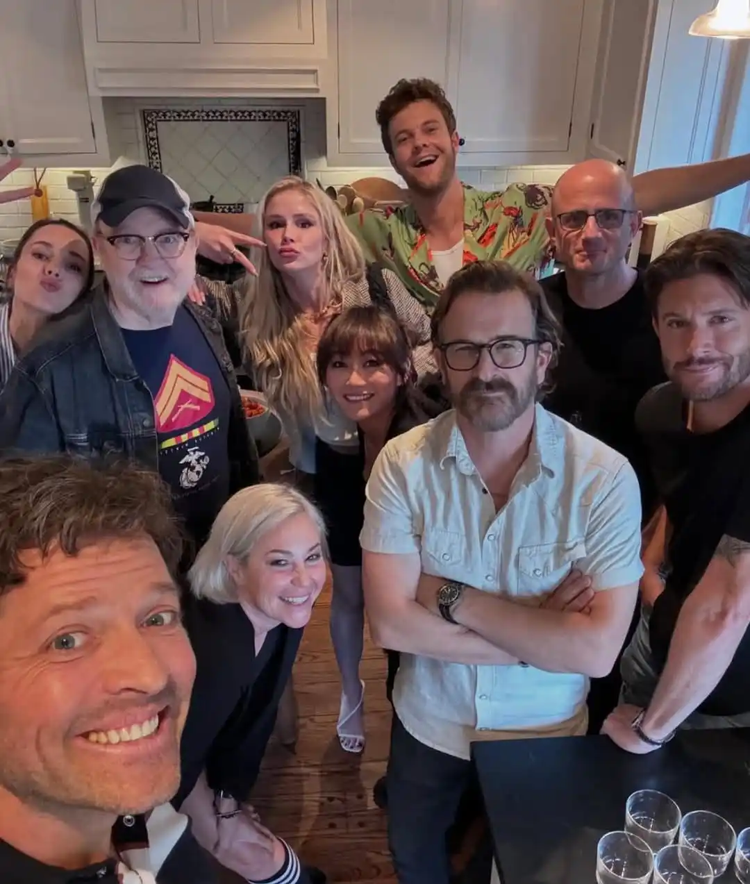 A selfie-style group photo of nine people in a kitchen, with the person taking the selfie in the foreground looking excited…