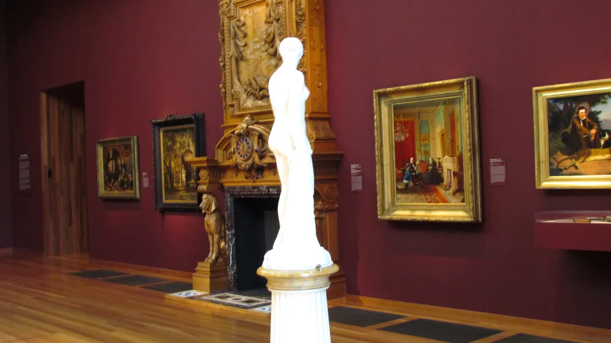 A white marble statue of a nude female figure stands on a pedestal in a museum gallery with deep red walls. Surrounding it…