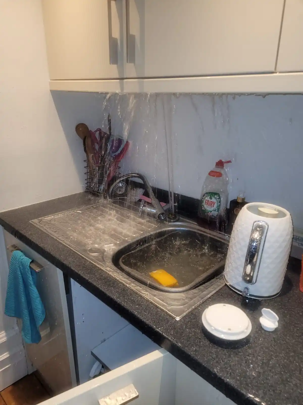 A kitchen sink with water spraying forcefully from the faucet, soaking the countertop and backsplash. A yellow sponge sits…