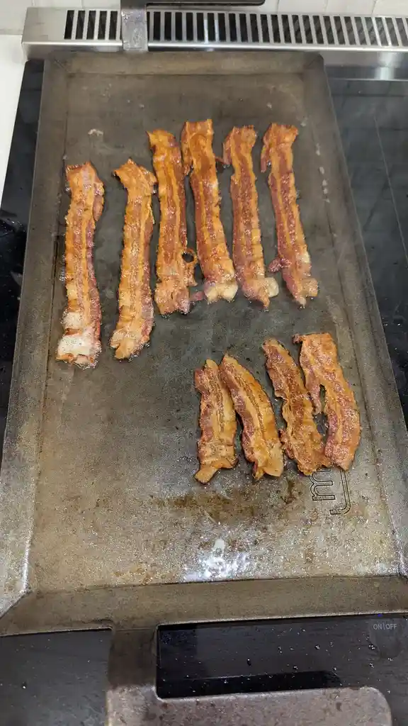 A close-up photo shows bacon strips sizzling on a metal griddle, with five longer strips arranged in a row on the top half…