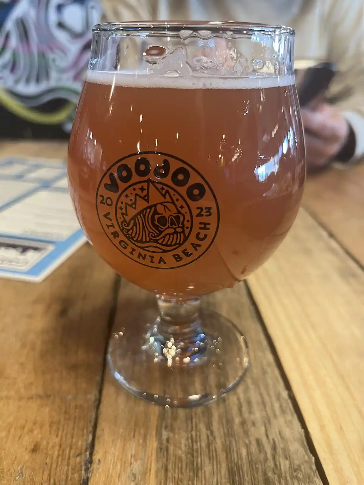 A close-up photo shows a stemmed beer glass filled with amber-colored beer and a white head, sitting on a wooden table. The…