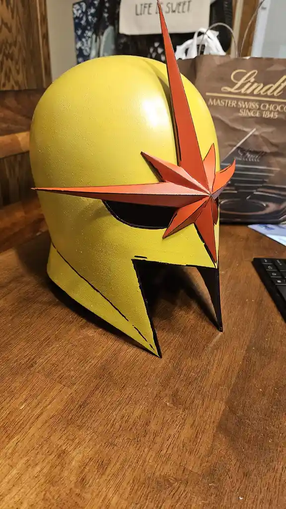 A yellow, star-adorned helmet resembling a superhero mask sits on a wooden table, with a Lindt chocolate bag and a 'Life is…
