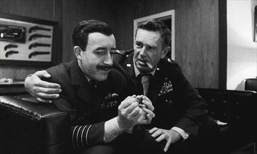 A black-and-white photo strip shows two men in military uniforms seated on a couch. The man on the left, with a mustache…