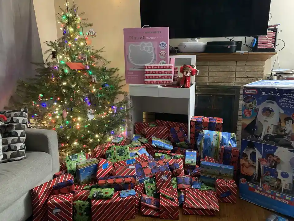 A living room is filled with a large pile of wrapped Christmas presents surrounding a decorated tree, with a Hello Kitty…