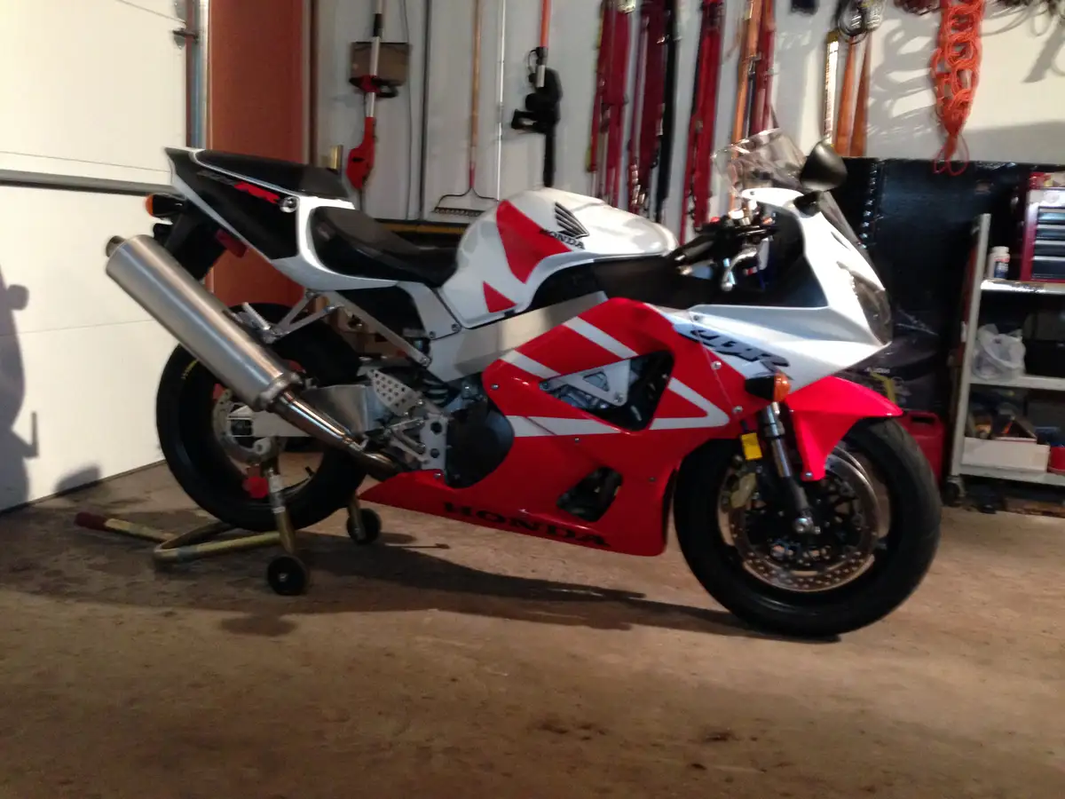 A red and white Honda sport motorcycle is parked on a stand in a garage, with tools and equipment visible on the walls…