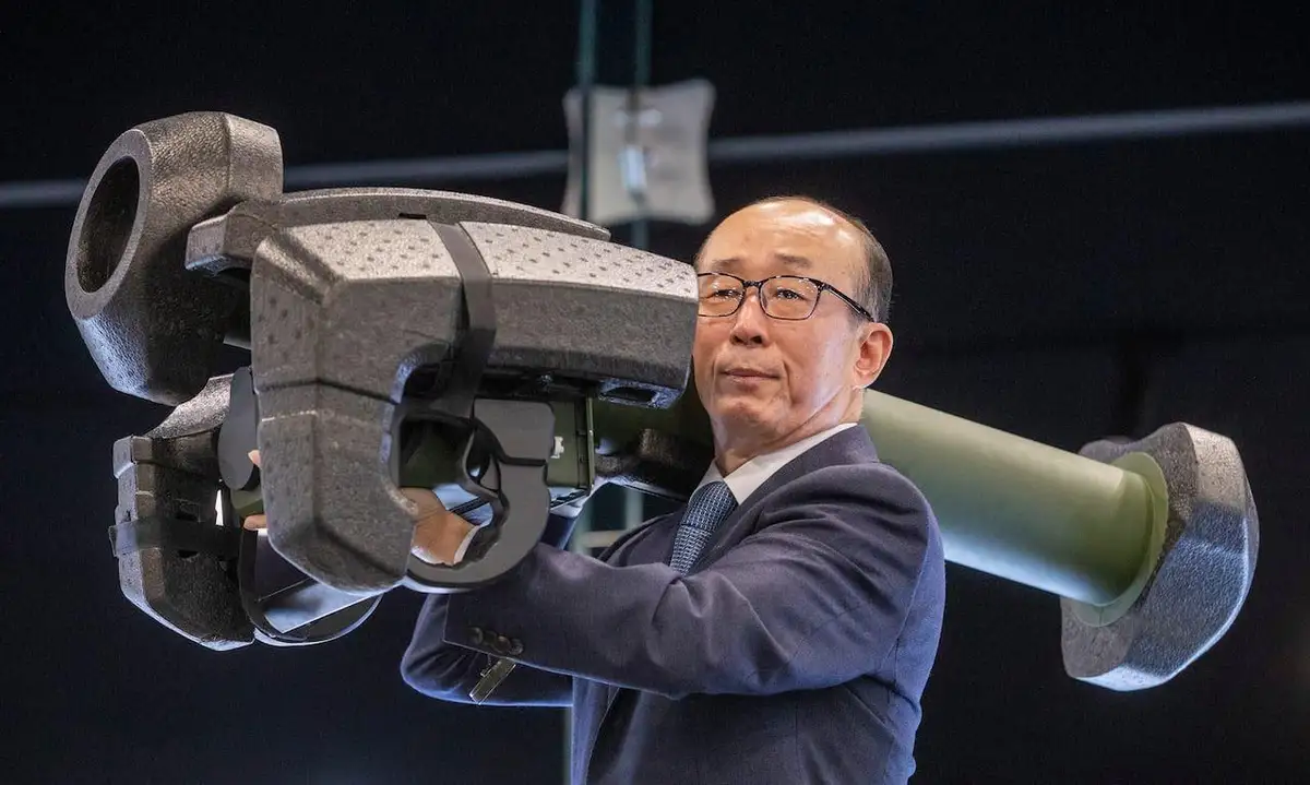 a man in a suit and glasses is holding a large, heavy-looking cannon-like object over his shoulder. The overlaid text reads…