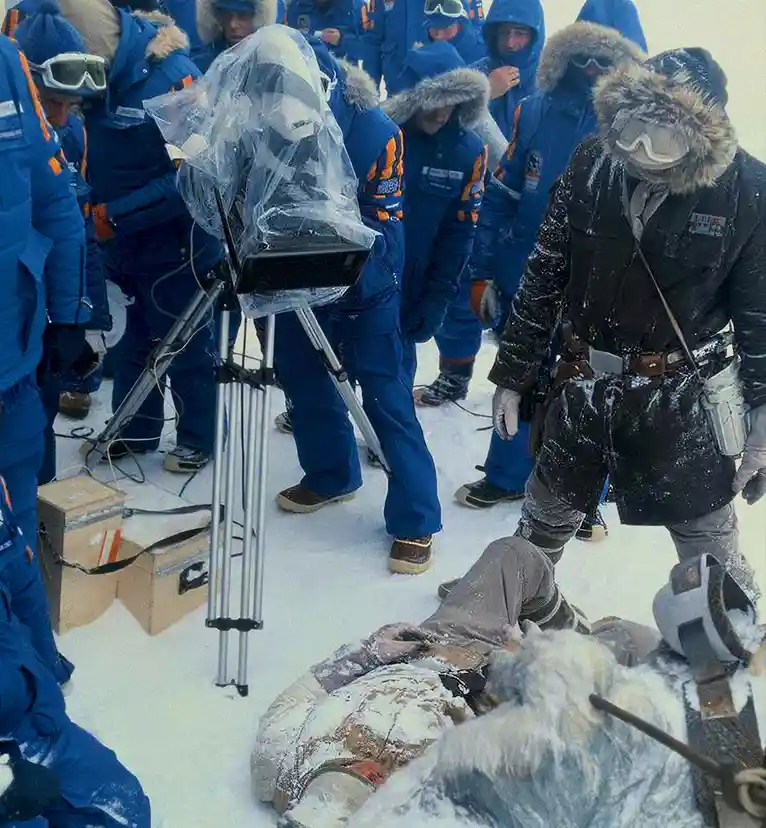 A group of people in heavy winter gear stand around a camera on a tripod in a snowy landscape. One person in a dark coat and…