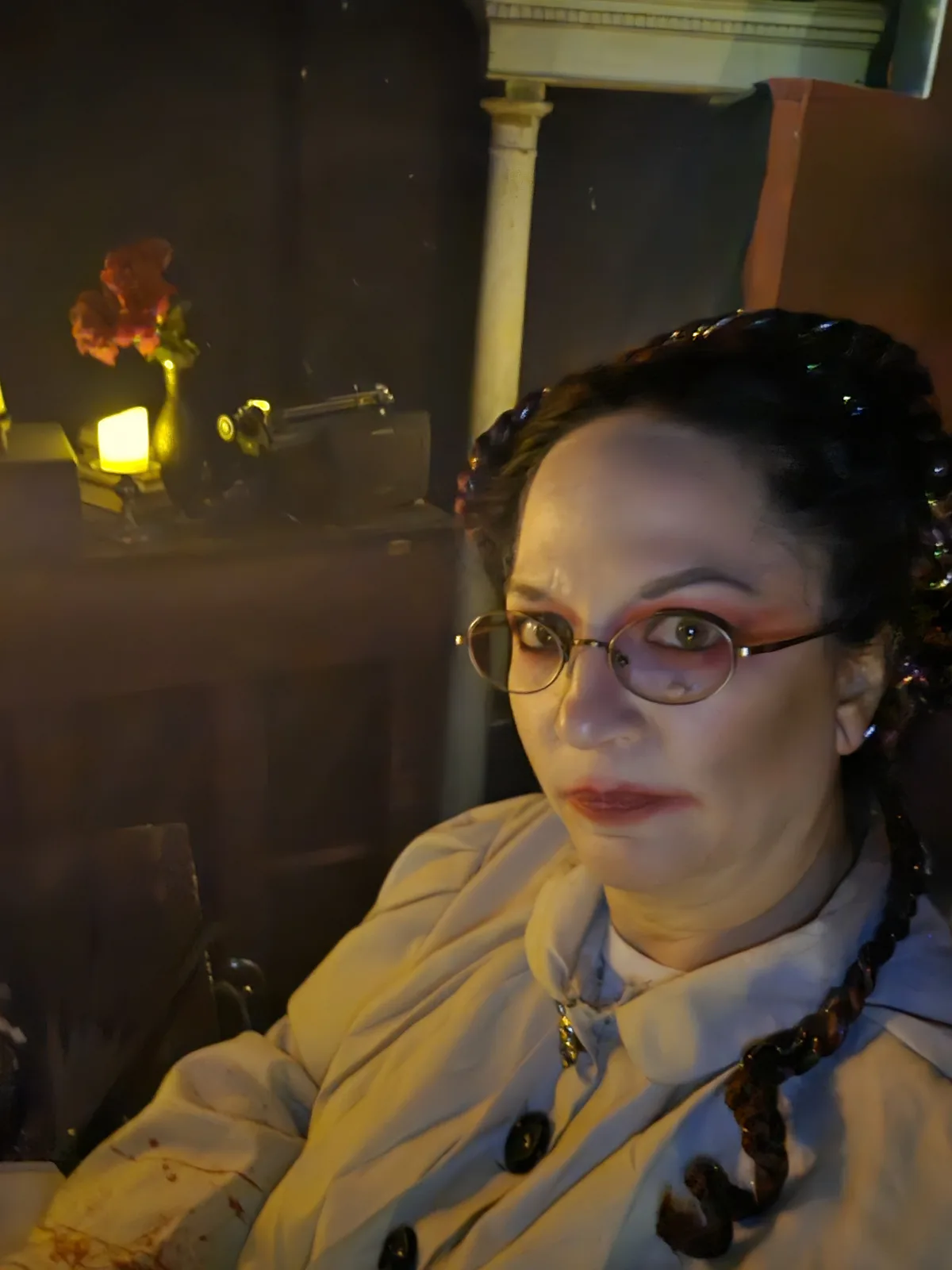 A person in a dimly lit room with vintage decor, wearing glasses, heavy makeup, and a light-colored coat with fake blood…