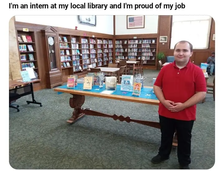 A man in a red polo shirt stands proudly in a library, smiling at the camera with his hands clasped. Behind him are wooden…