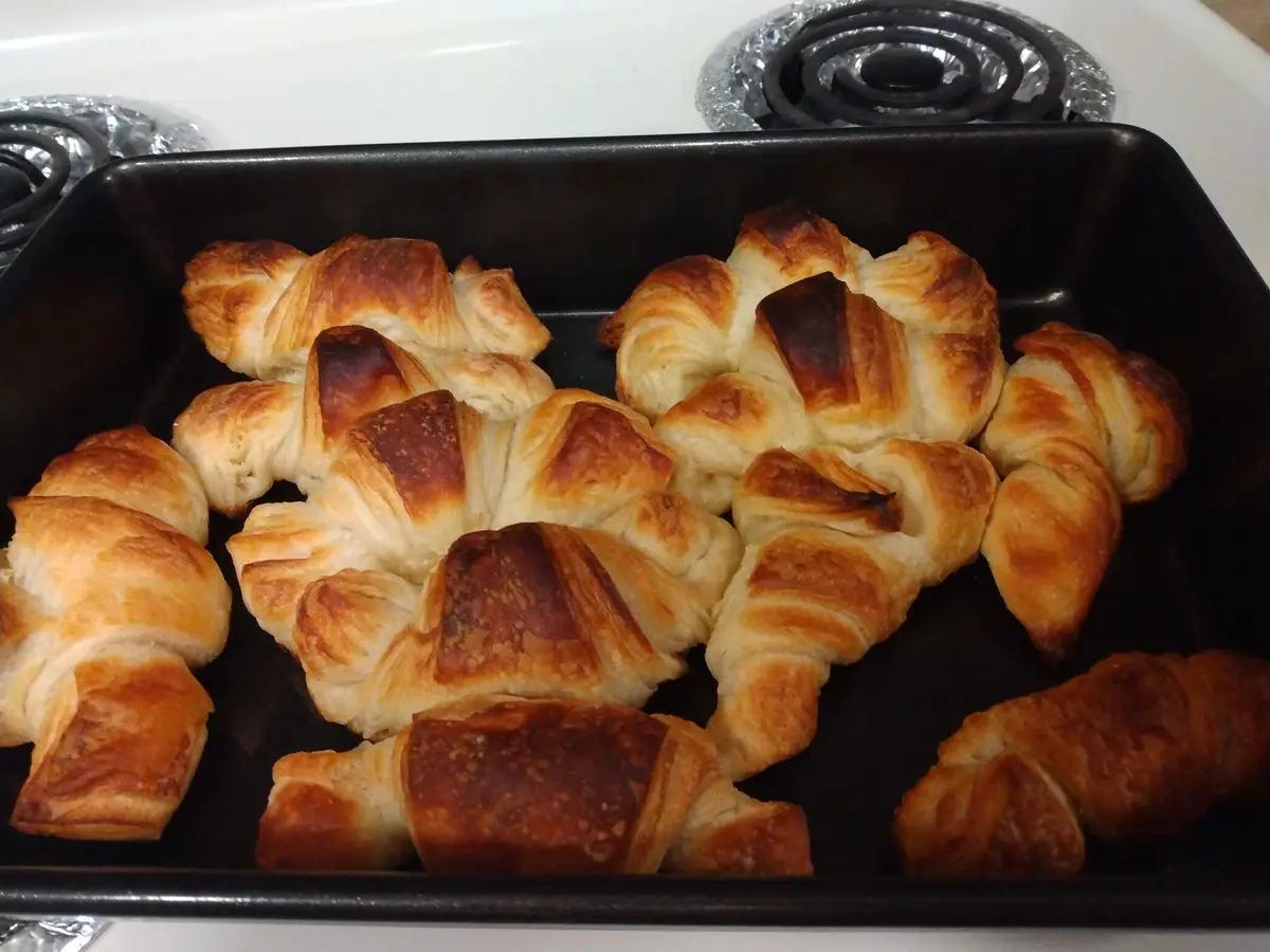 A black baking tray holds several golden-brown croissants, some of which are visibly burnt on top, sitting on a white…