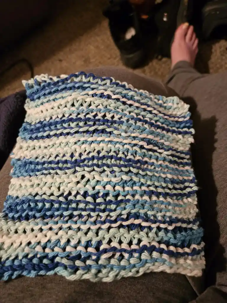 A close-up photo shows a hand-knitted textile with horizontal stripes of blue, light blue, and white yarn, resting on…
