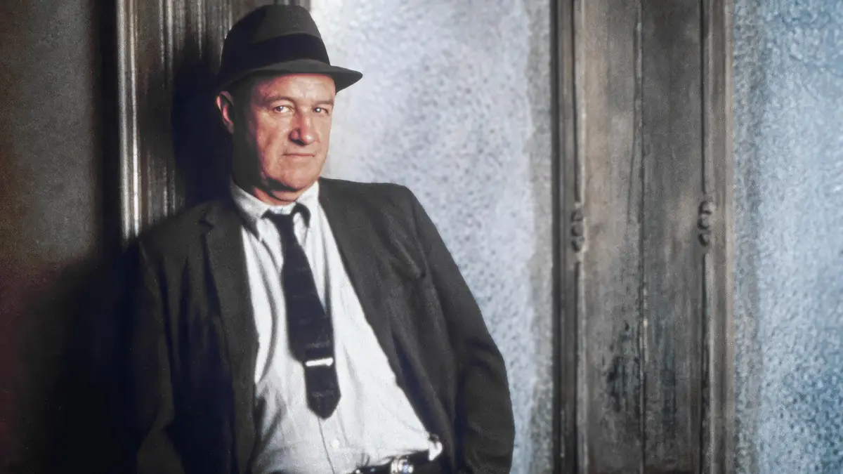 a man in a dark suit, white shirt, dark tie, and fedora leans against a textured wall, looking directly at the camera with a…