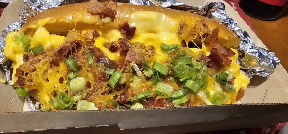 A close-up photo of a hot dog in a cardboard tray lined with foil, generously covered in melted yellow cheese, crispy bacon…